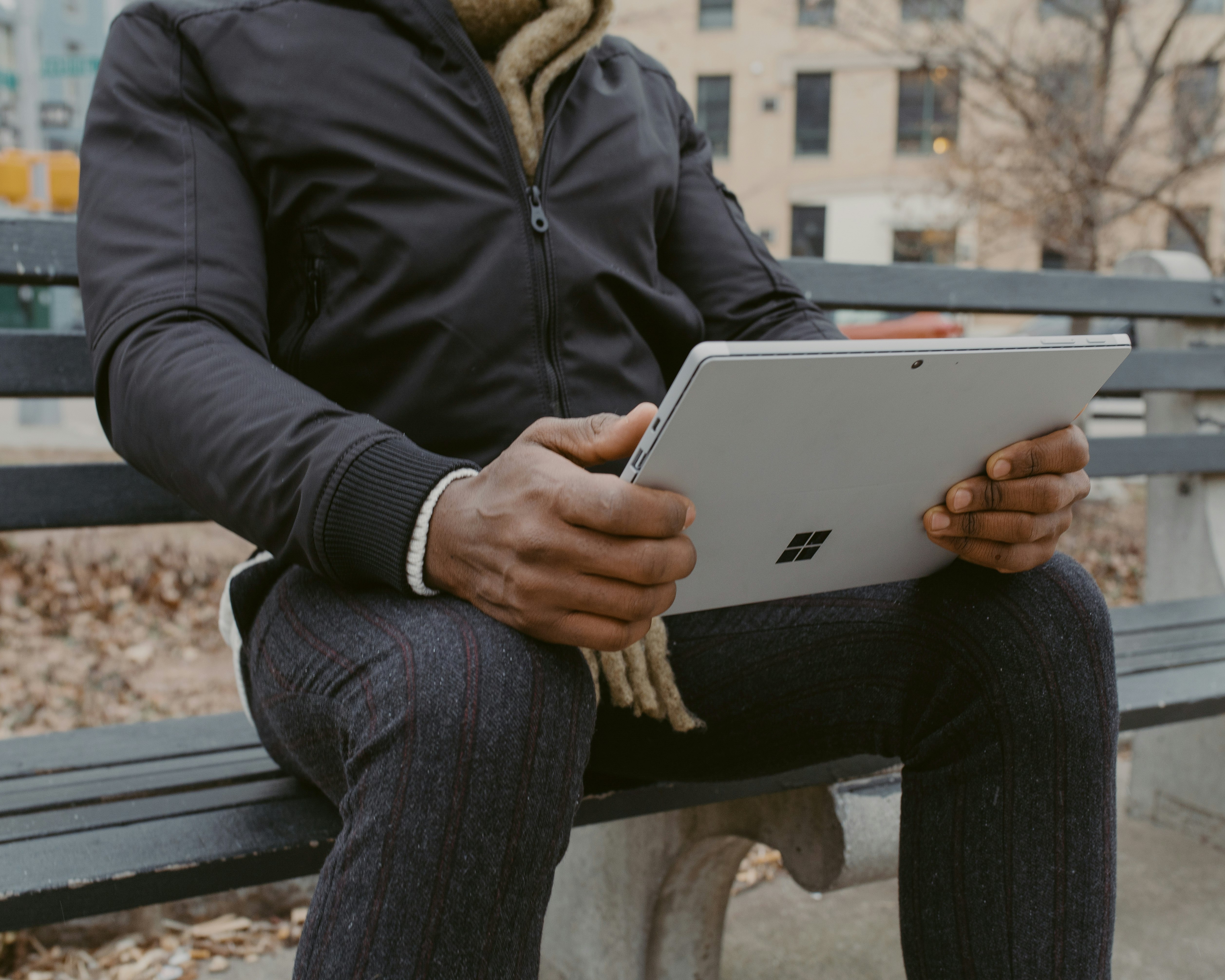 person in black jacket and gray pants sitting on bench holding silver Surface